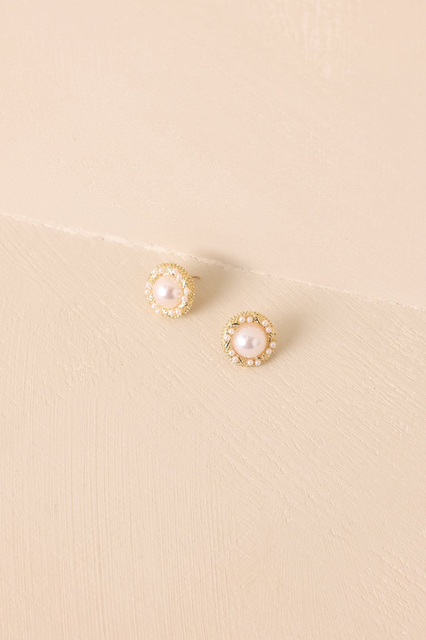 Cropped view of the stud earrings with round faux pearl center framed by a textured gold border and small pearls.