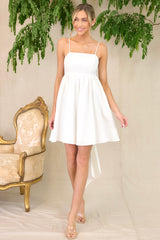 Full body view of model walking in this dress that features a square neckline, skinny adjustable shoulder straps, a back zipper with a wrap around self-tie bow, waist pockets, and flowy short skirt