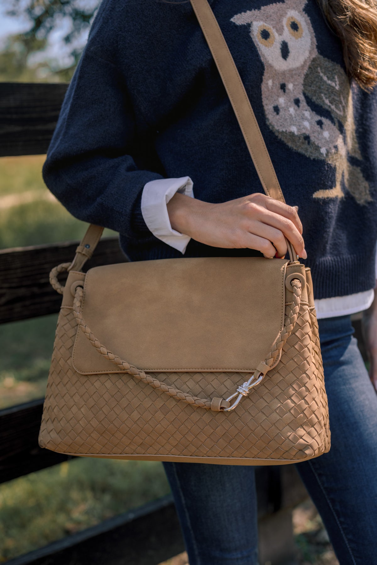 Tan basketweave handbag featuring a smooth flap top, braided handles, and silver-tone decorative accent.