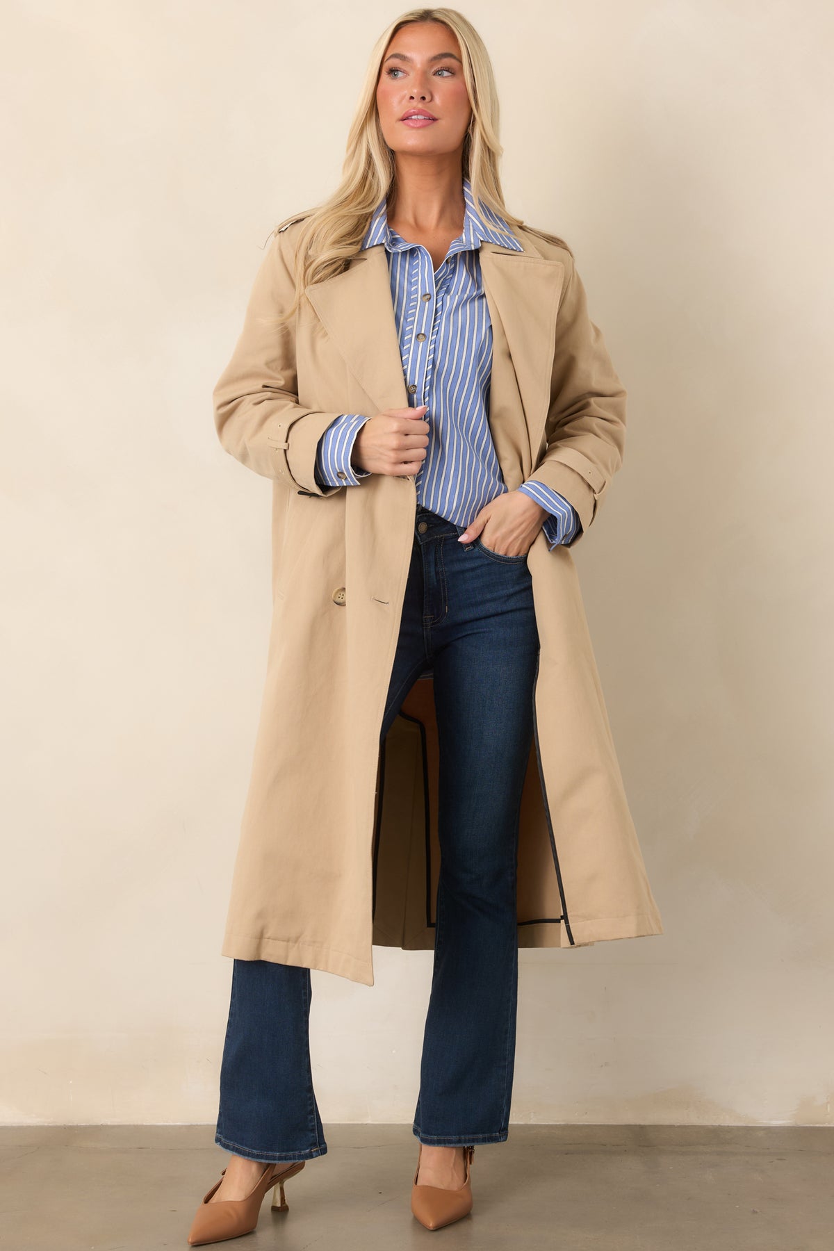 Tan trench coat with a refined double-breasted design, defined waist belt, functional pockets, and flowing full-length construction.