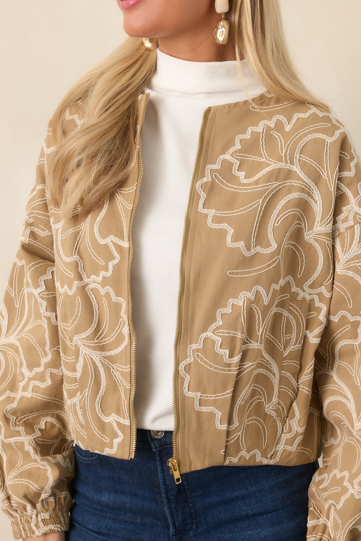 Close-up of the ivory floral embroidery and gold-tone zipper hardware against the tan cotton fabric.