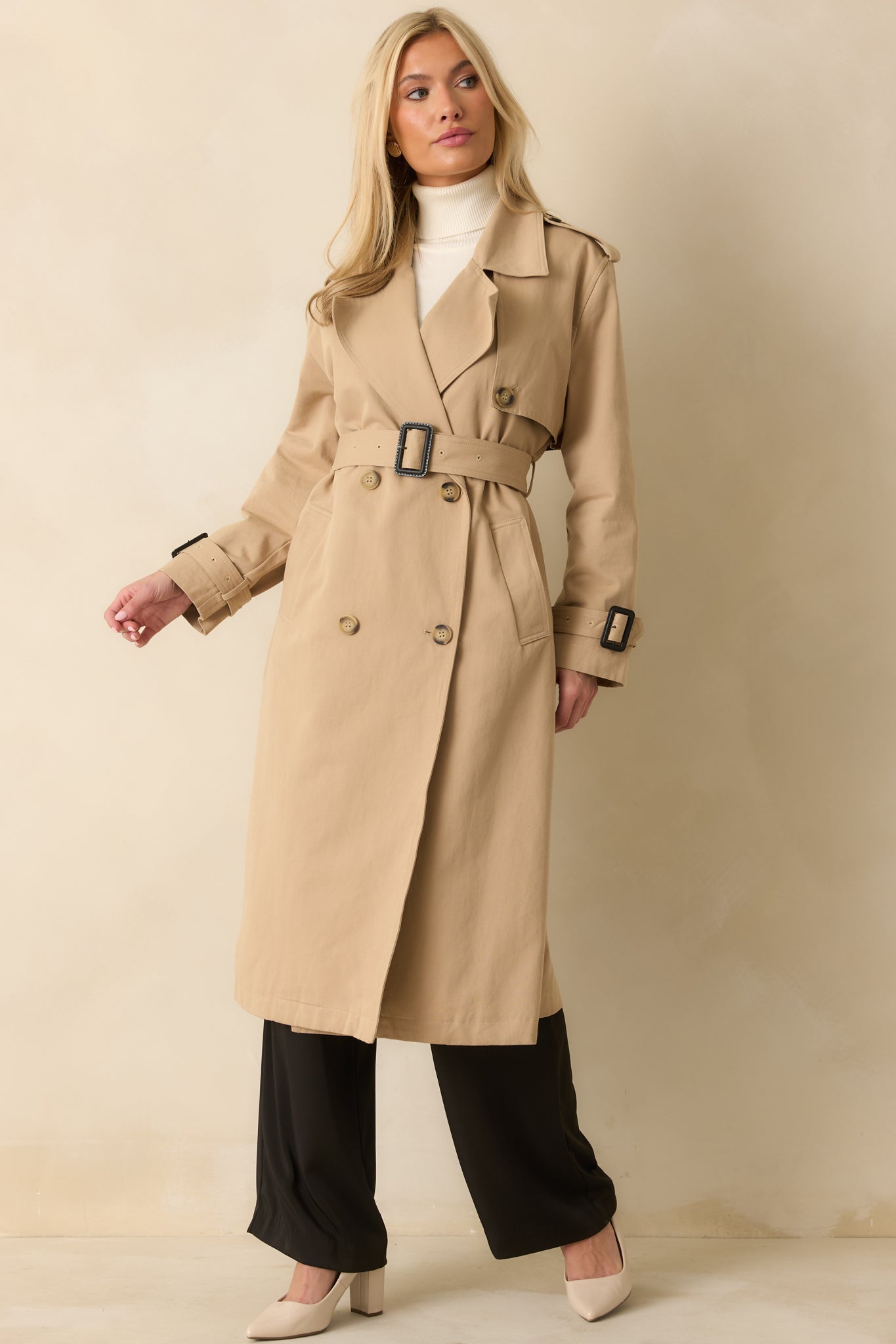 Front view of the tan trench coat highlighting storm flap, adjustable buckled cuffs, and polished silhouette.