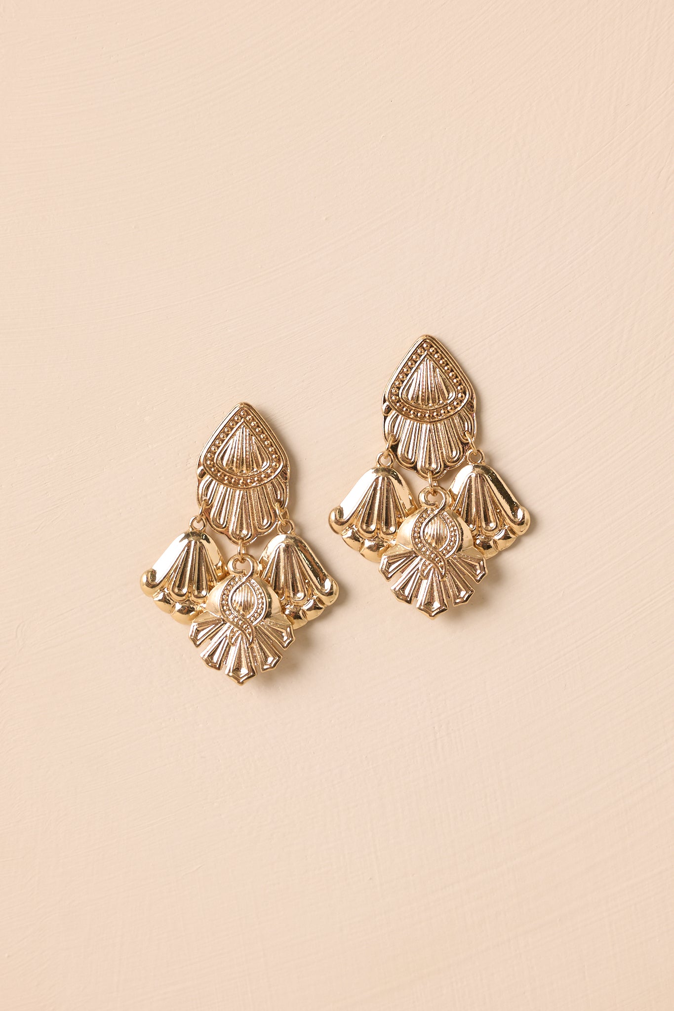 A shot of the textured gold chandelier earrings with three drop charms shown against a beige background.
