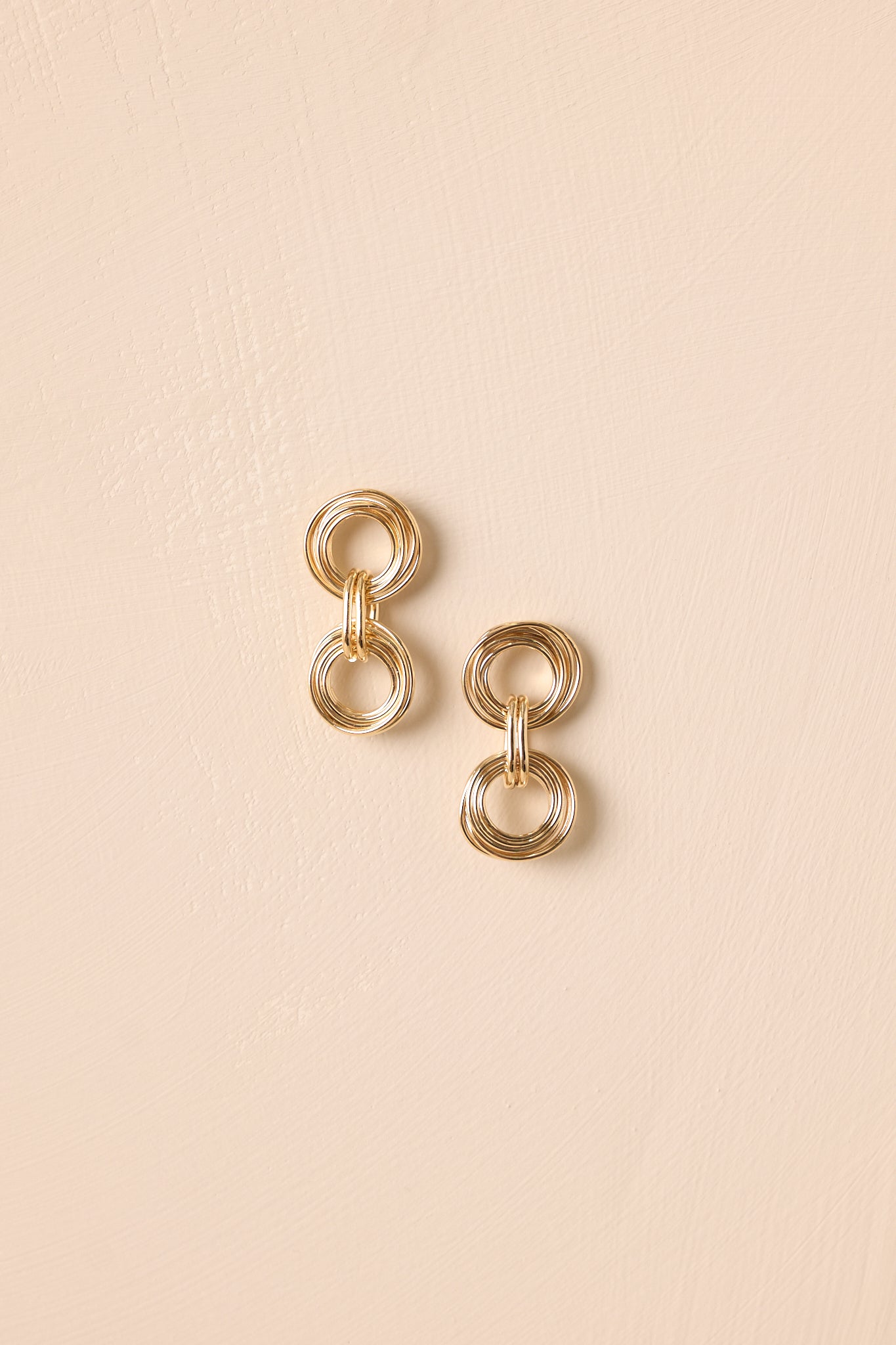 A shot of the textured gold drop earrings with layered circular links shown against a beige background.