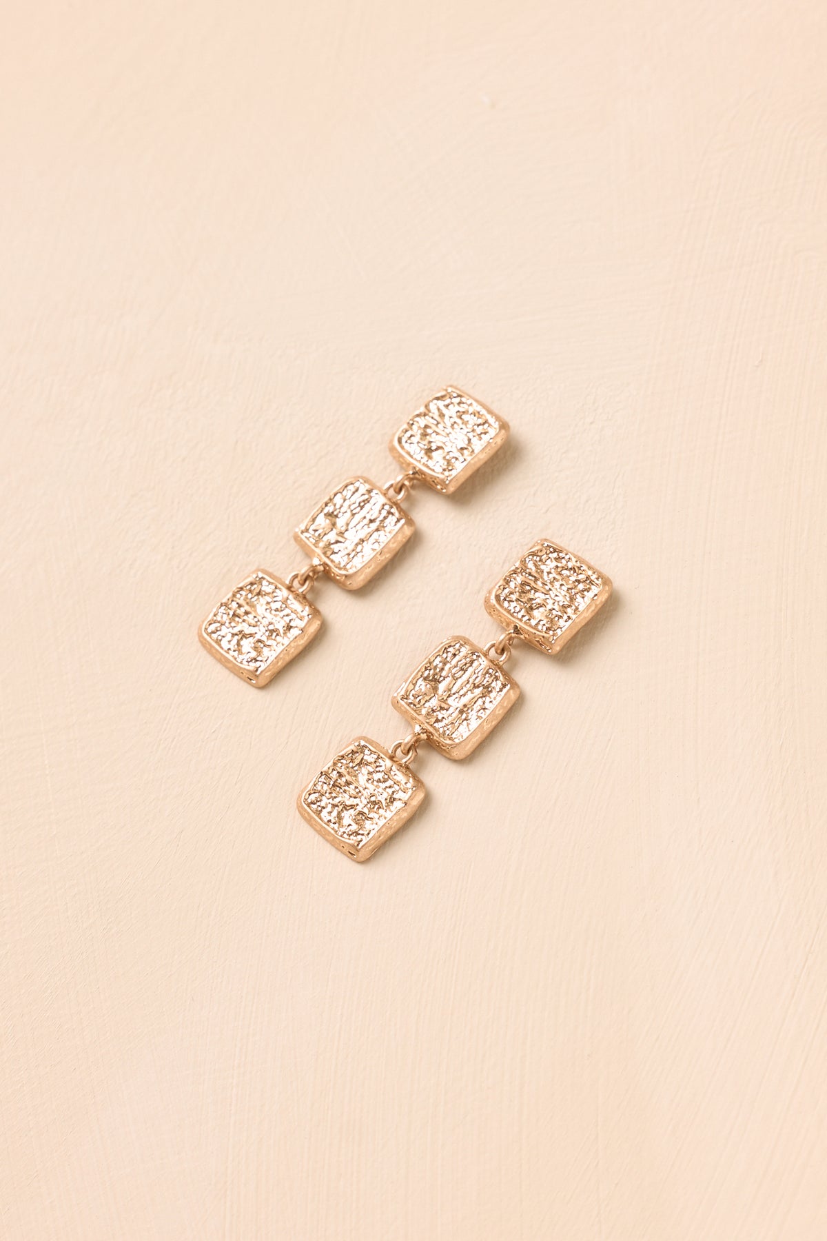 Textured square earrings displayed against a beige background, featuring three linked geometric shapes with a sculptural silhouette.