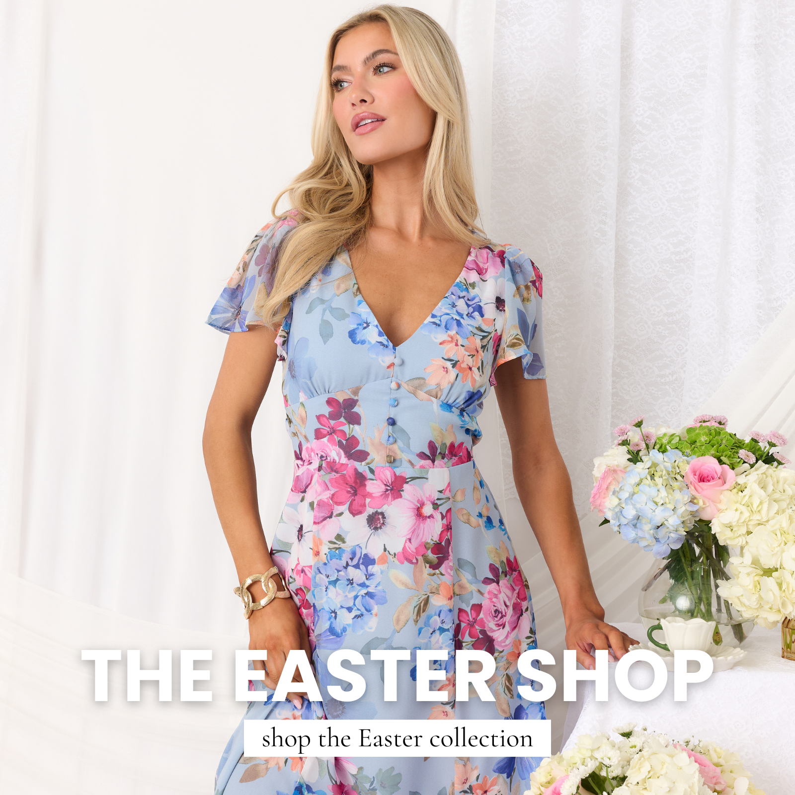 Model wearing a light blue floral v-neck flutter sleeve dress standing beside pastel flowers and lace draping, promoting The Easter Shop collection.