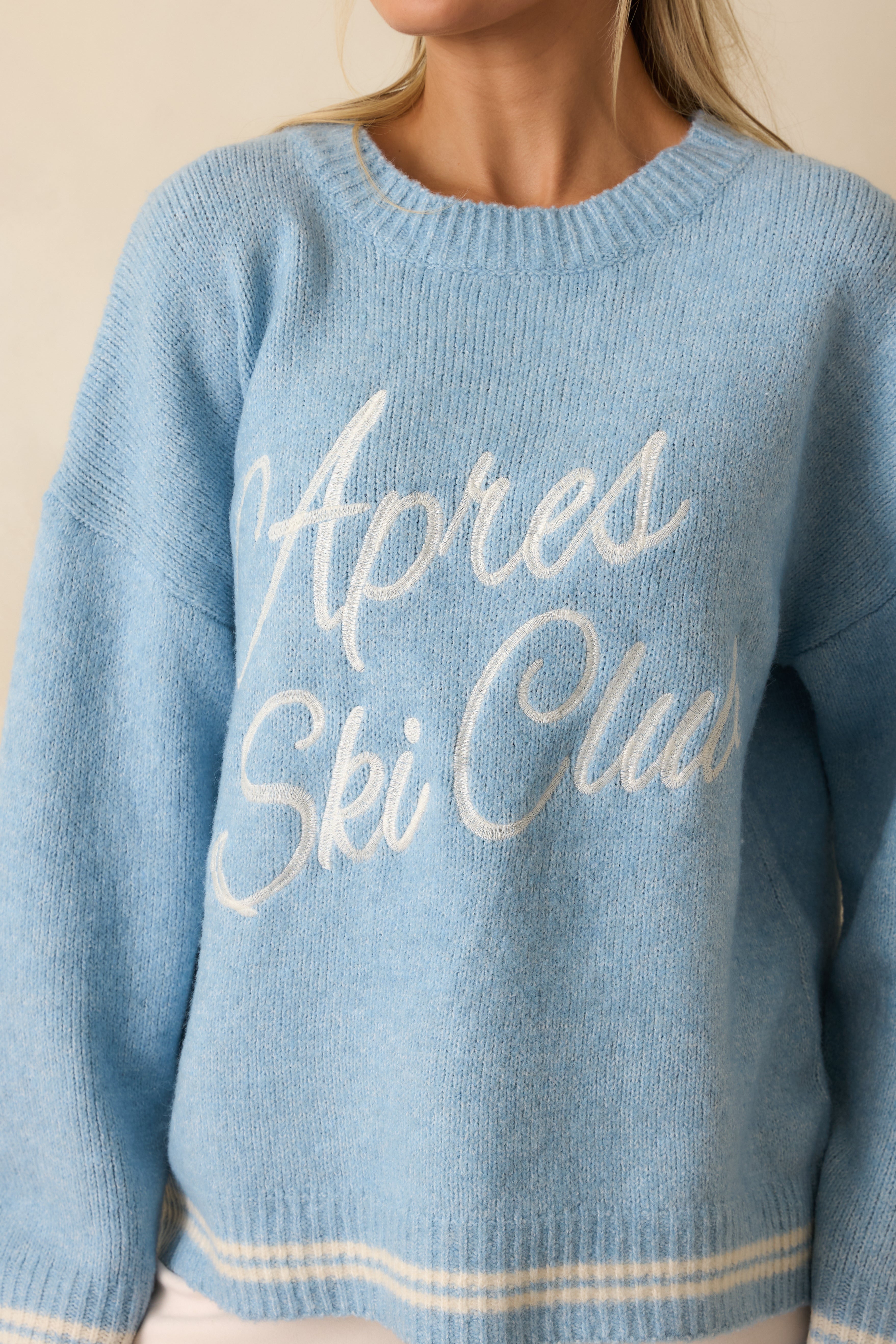 Tight view of white “Après Ski Club” embroidery on powder blue knit fabric, showing ribbed hem and texture.