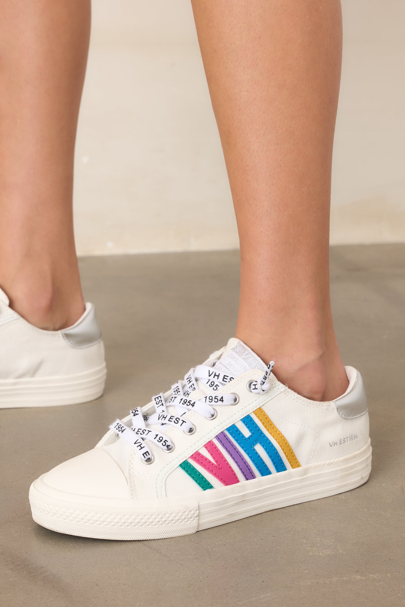 White canvas low-top sneakers displayed against a beige background, featuring colorful retro “VH” side detailing, silver heel accents, and lace-up closures.