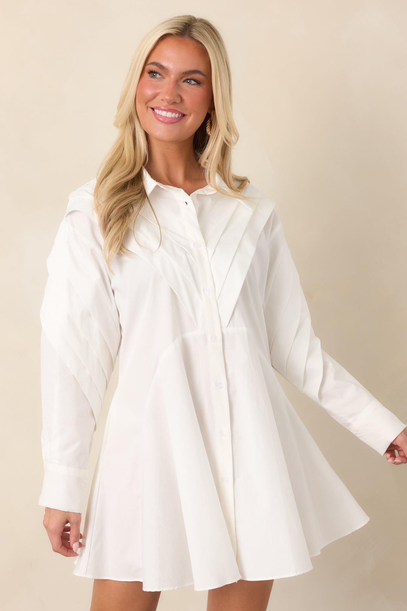 Close view of the white mini dress highlighting the collared neckline, button-front closure, and layered pleated detailing across the shoulders.