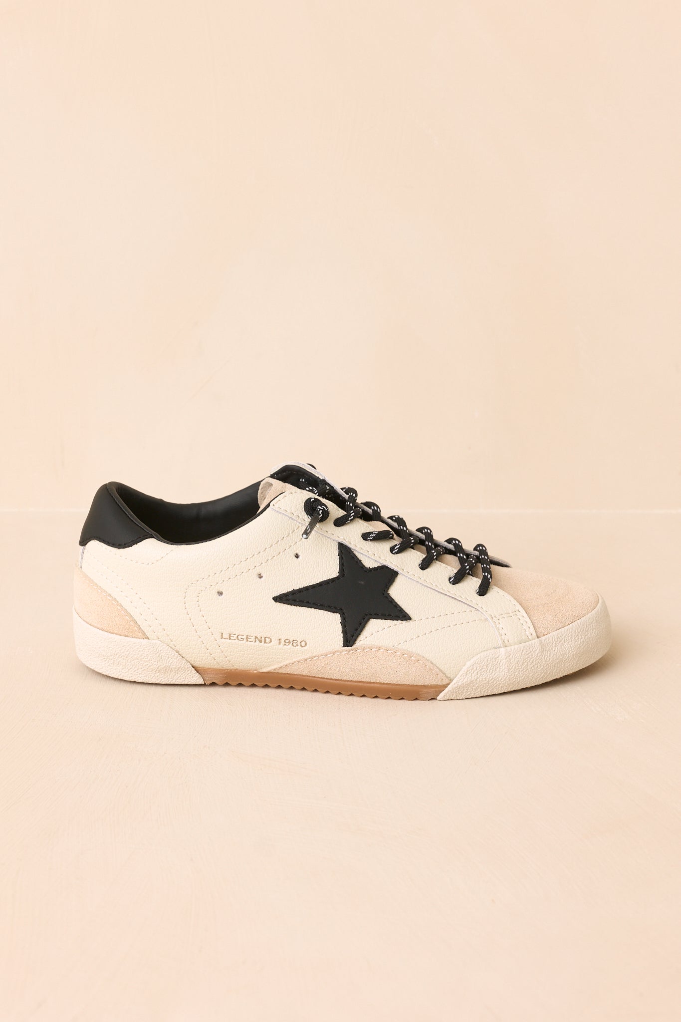 White low-profile sneakers featuring a bold black star, black heel accent, textured sole, and a vintage-inspired worn finish displayed against a beige background.
