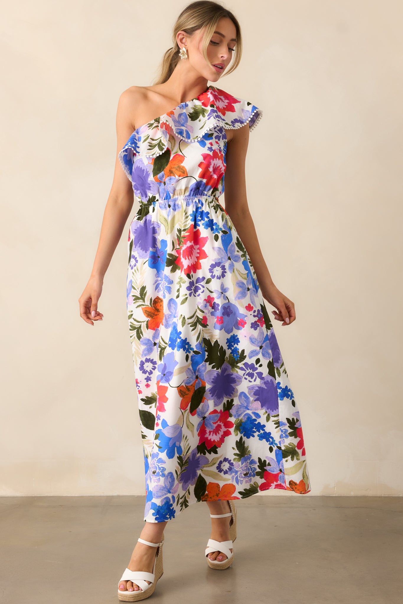 A front view of the white maxi dress featuring a blue and red floral print, an elastic one-shoulder neckline with ruffles, and a comfortable elastic waist, paired with functional pockets for practicality.