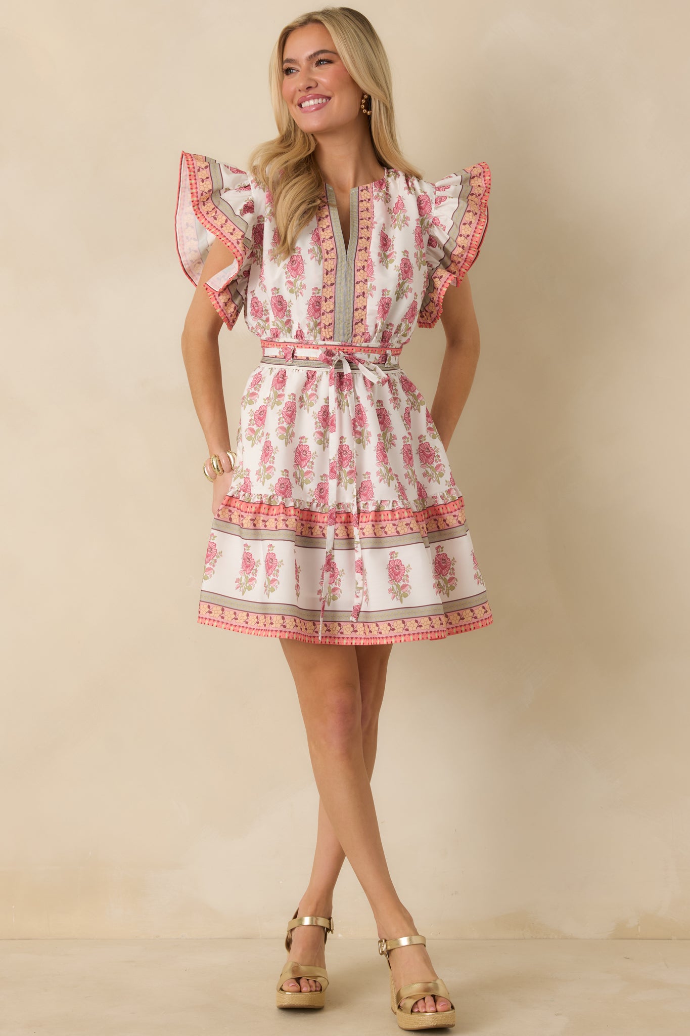 White mini dress with an allover floral print, V-neckline, flutter sleeves, and tiered hem accented with orange and green trim.