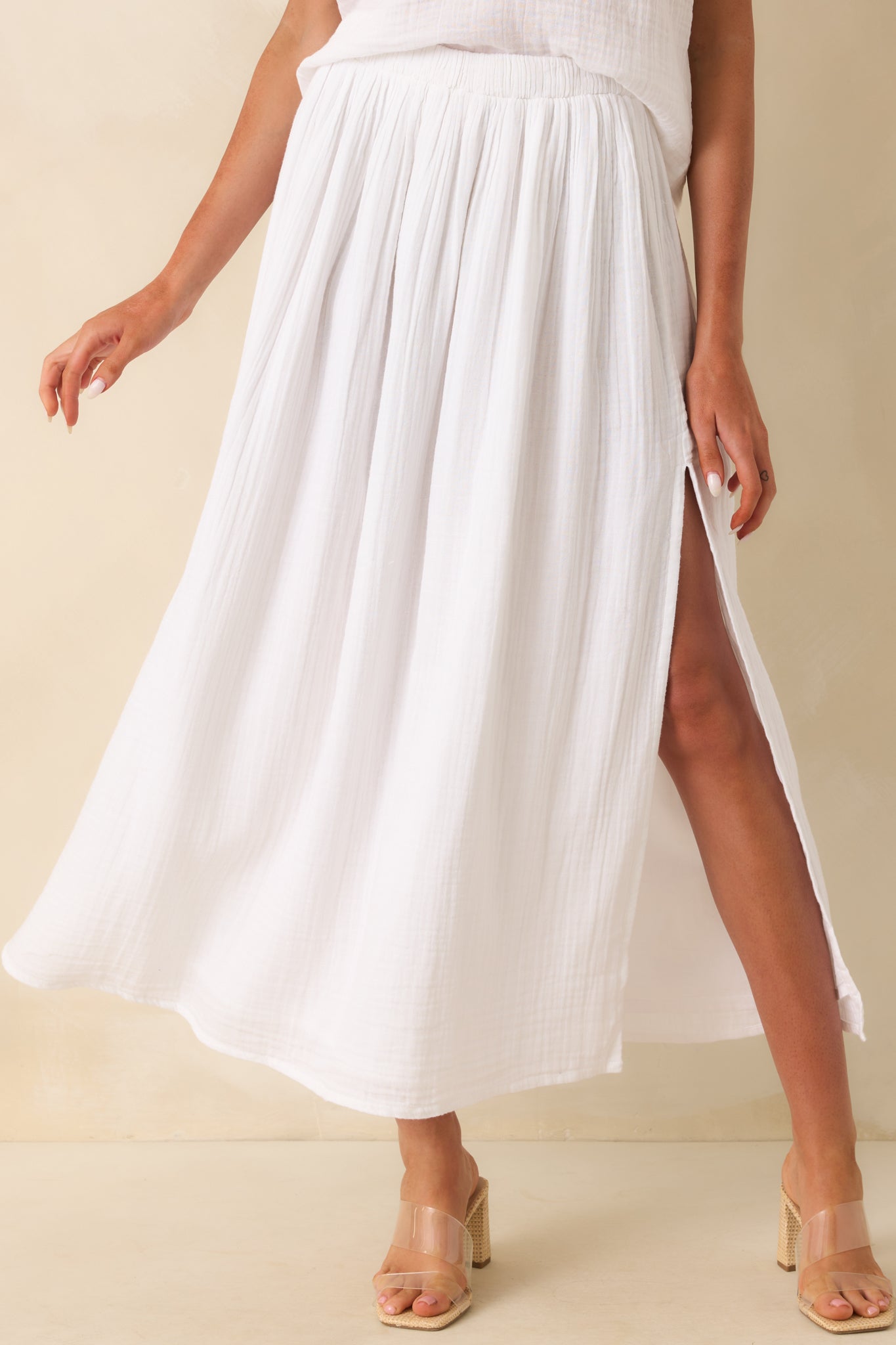 Up close photo of model wearing a white skirt that features a lightweight 100% cotton gauze fabric, high rise design, elastic waistband, relaxed fit, midi length, and leg slit.