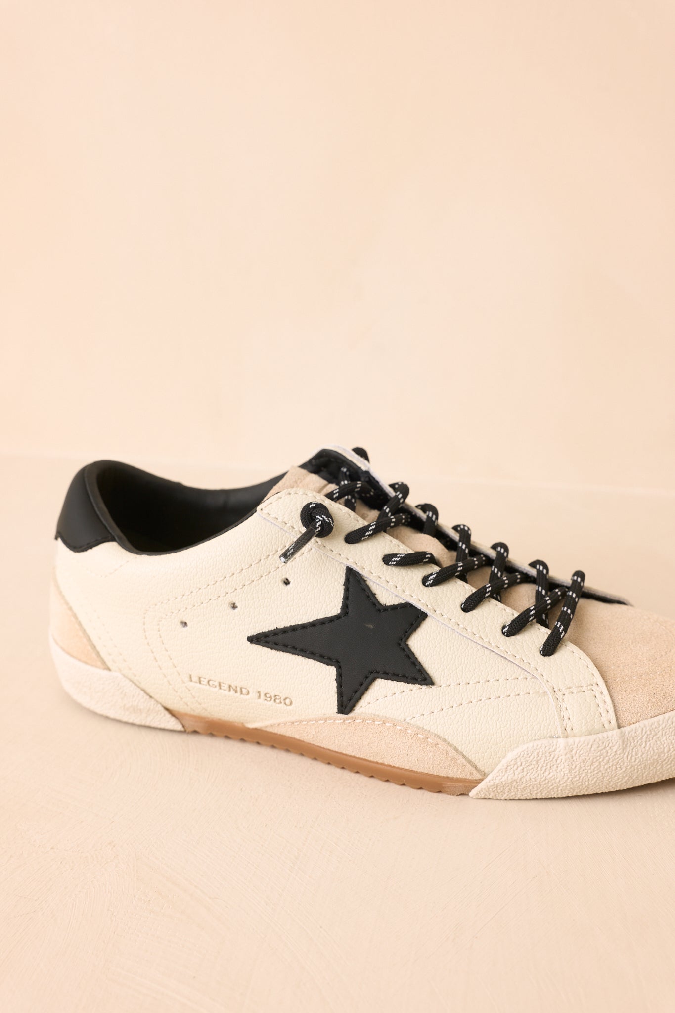 Detailed view of the white sneaker emphasizing the black heel accent, textured sole, mixed fabrics, and vintage-inspired styling on a beige background.