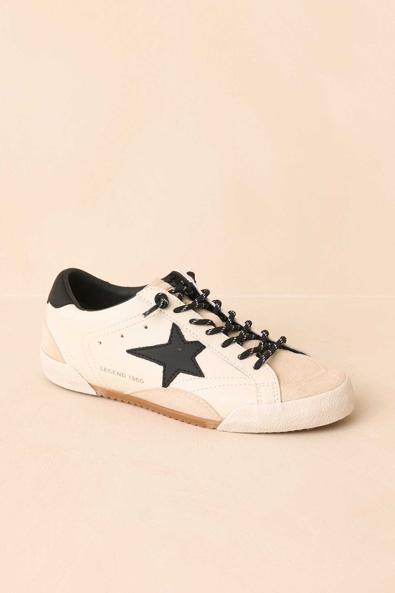 White sneaker highlighting the bold black star, layered material construction, precise stitching, and lightly distressed finish against a beige background.