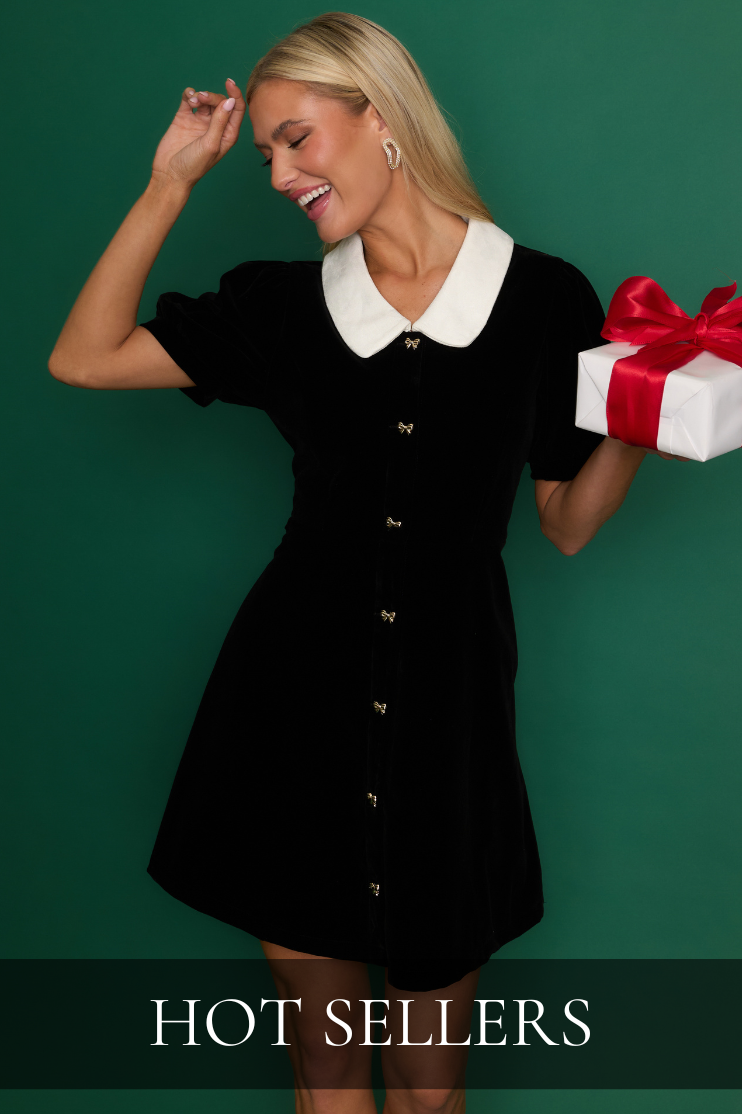 Woman wearing a black velvet mini dress with a white collar and gold buttons, holding a gift box—classic holiday party look for women’s fashion. Outfit is featured in our Hot Sellers collection.