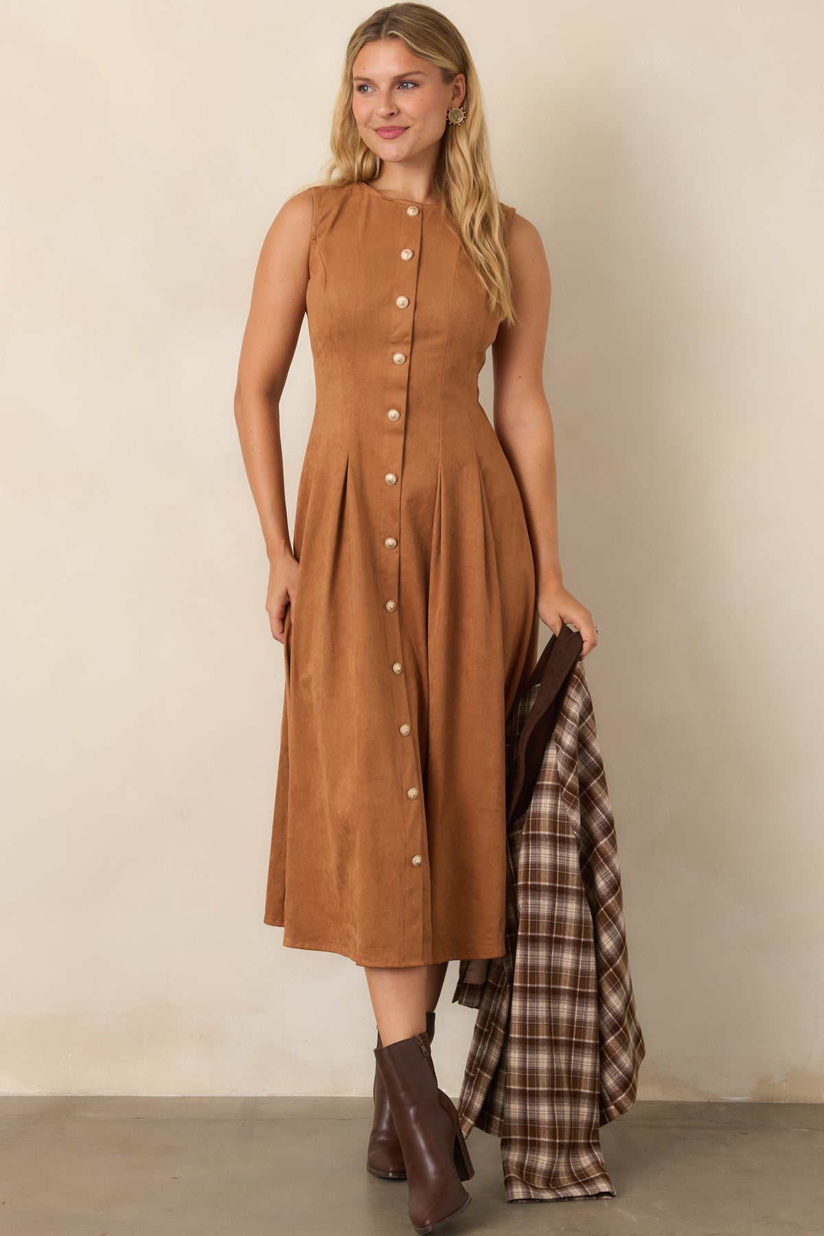 A woman models a sleeveless, midi-length brown dress with a button-down front, holding a brown plaid shirt jacket. She is wearing brown leather ankle boots against a neutral beige studio background.