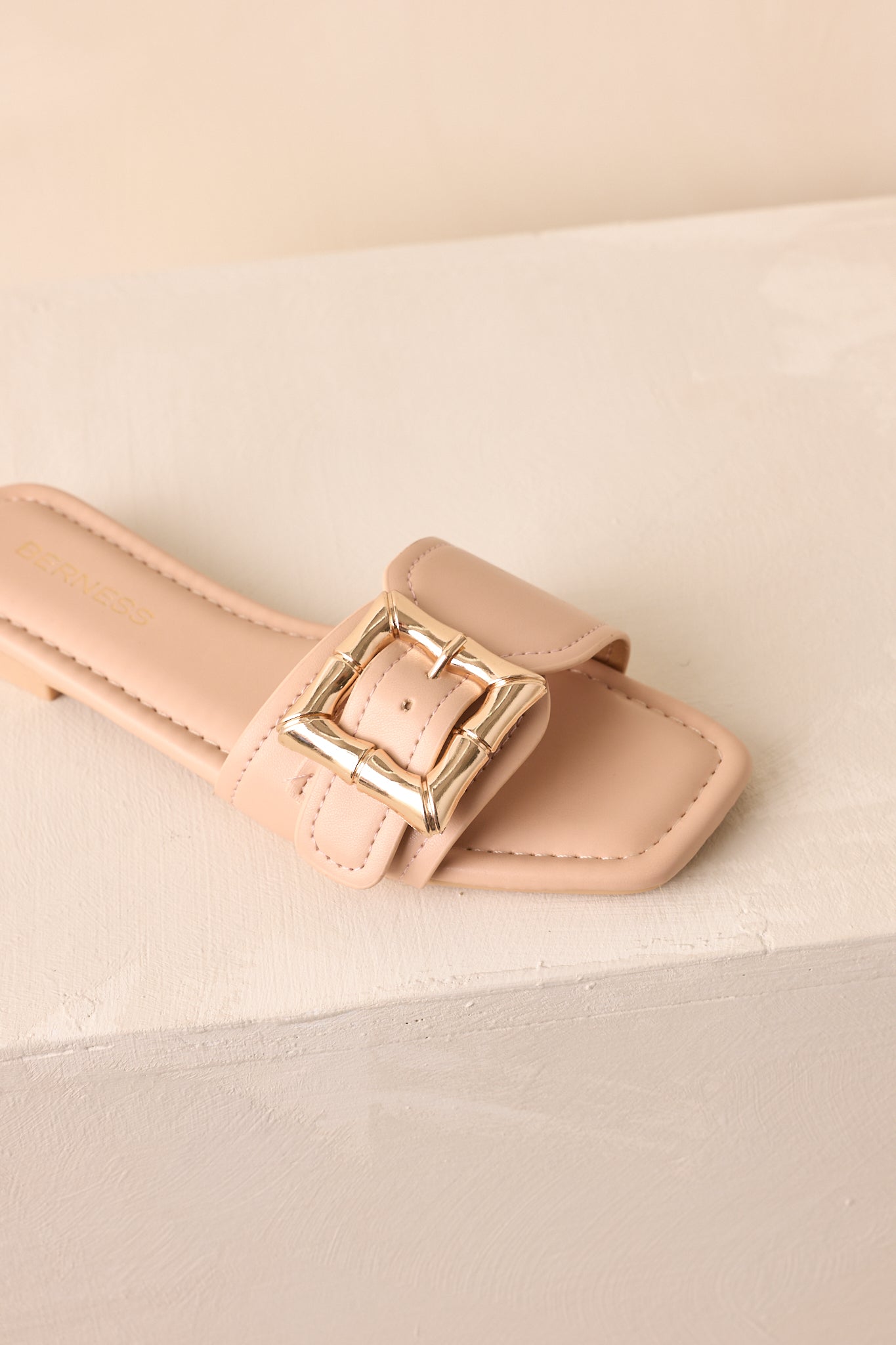A zoomed-in front view of the beige sandal emphasizing its gold buckle and open-toe design.
