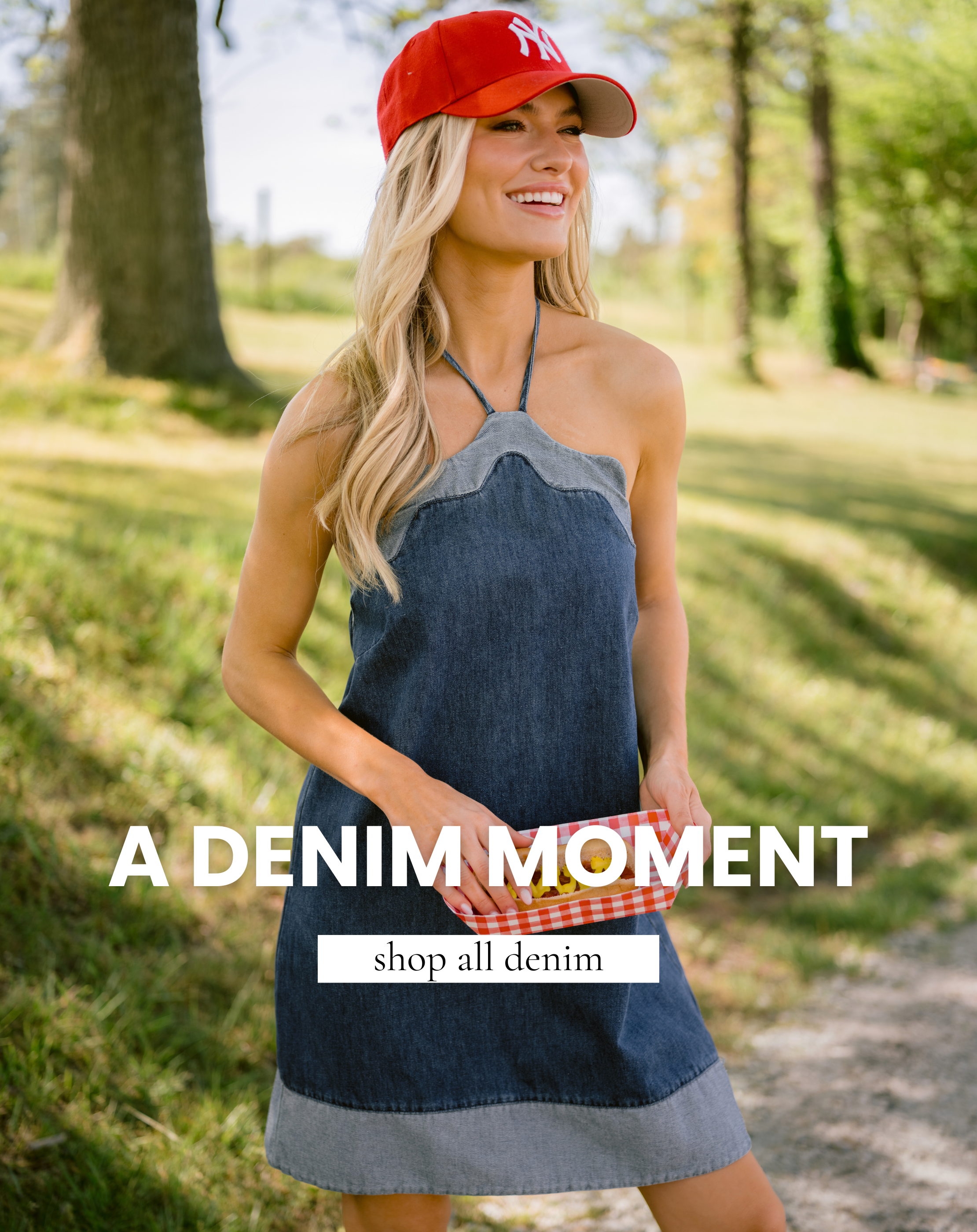 Model wearing a dark wash denim mini dress with a halter neckline and a red hat, standing in a green field. Dress is featured in the All Denim collection.