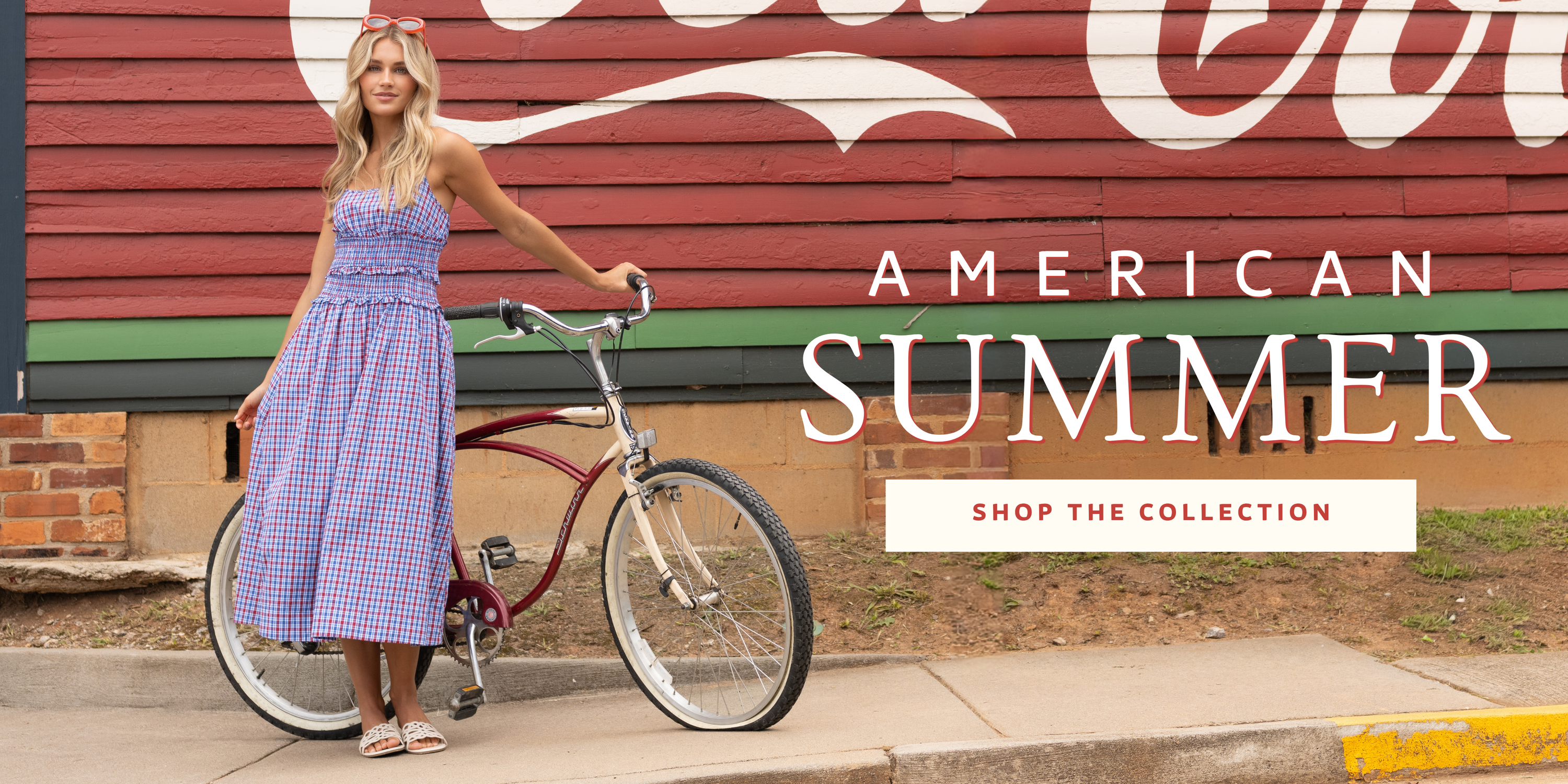 Woman wearing a red, white, and blue gingham smocked midi dress standing with a vintage bicycle in front of a painted American-themed mural promoting the American Summer collection.