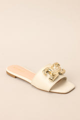 Front angled view of these ivory slip-on sandals that are adorned with a striking large gold buckle-like design, adding a touch of glamour to your ensemble.