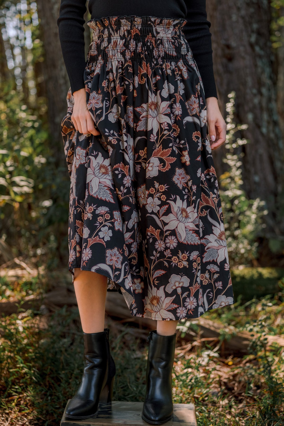 Model wearing a black floral midi skirt with a smocked waist, styled with a black sweater and black ankle boots.