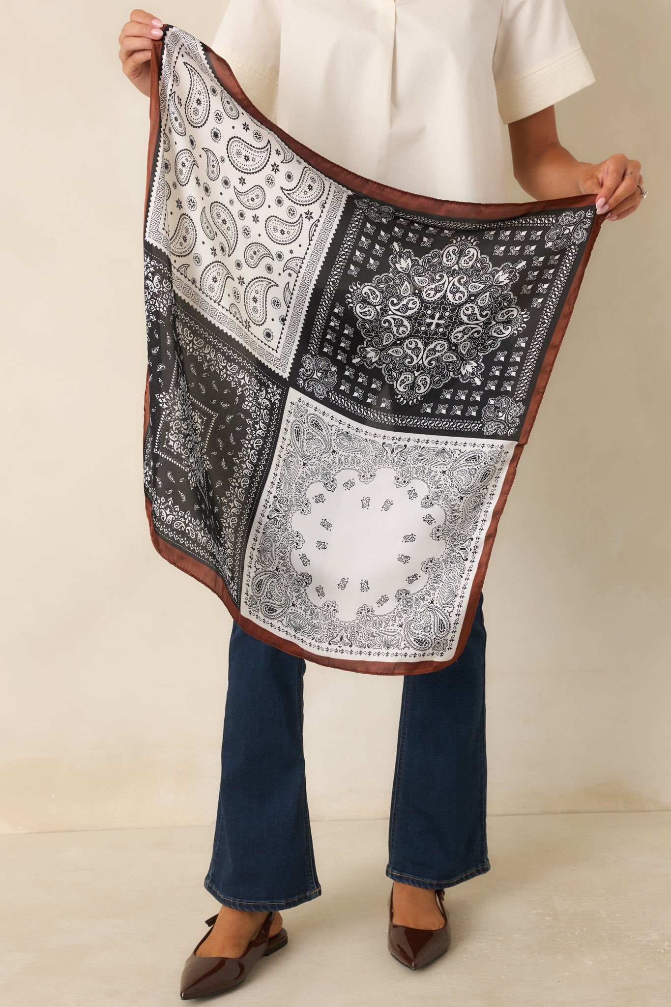 Model holding a brown, black, and ivory square bandana scarf featuring a patchwork paisley print and contrasting brown border.