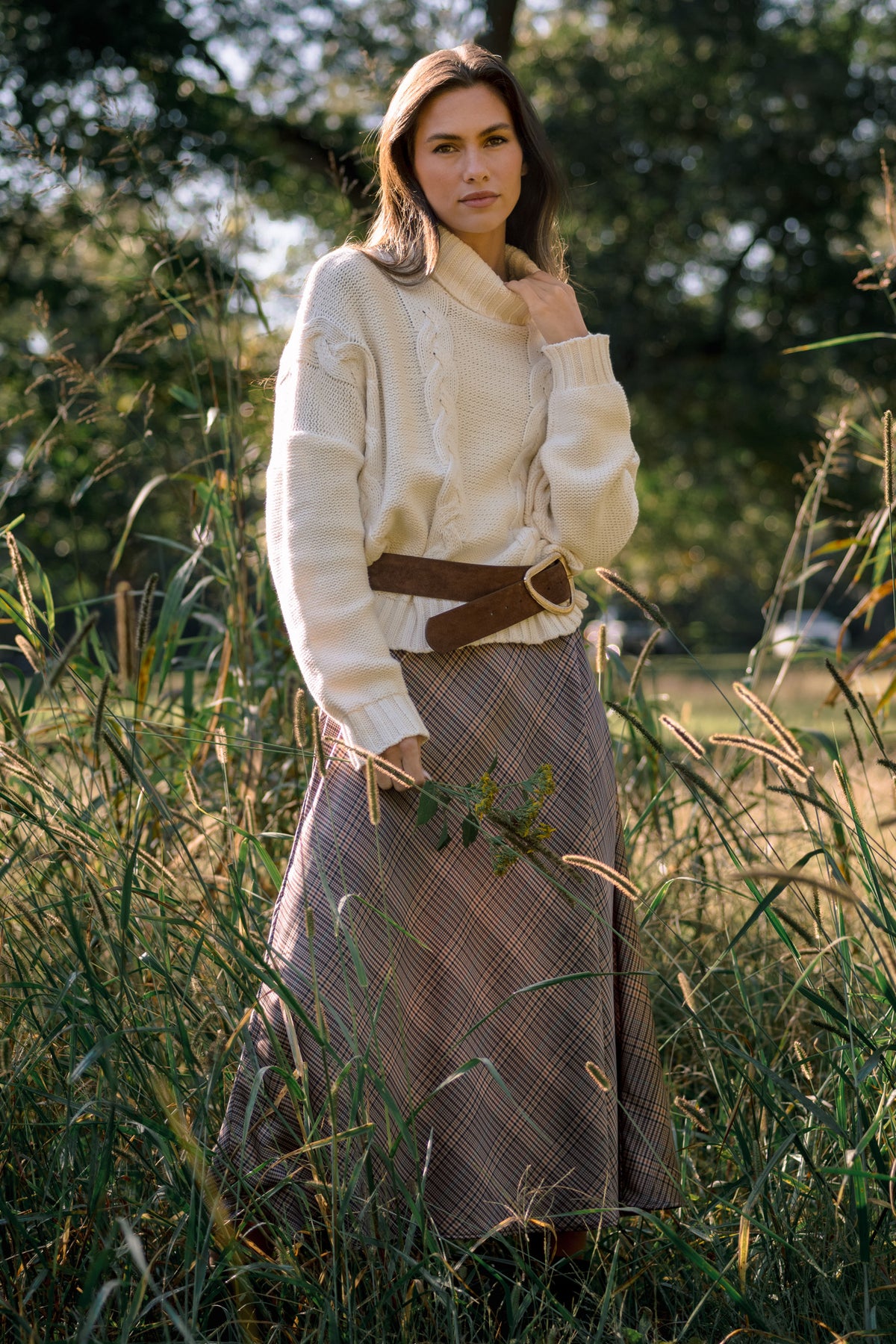 Model wearing a brown plaid maxi dress styled with a cream cable-knit sweater and wide brown belt outdoors.