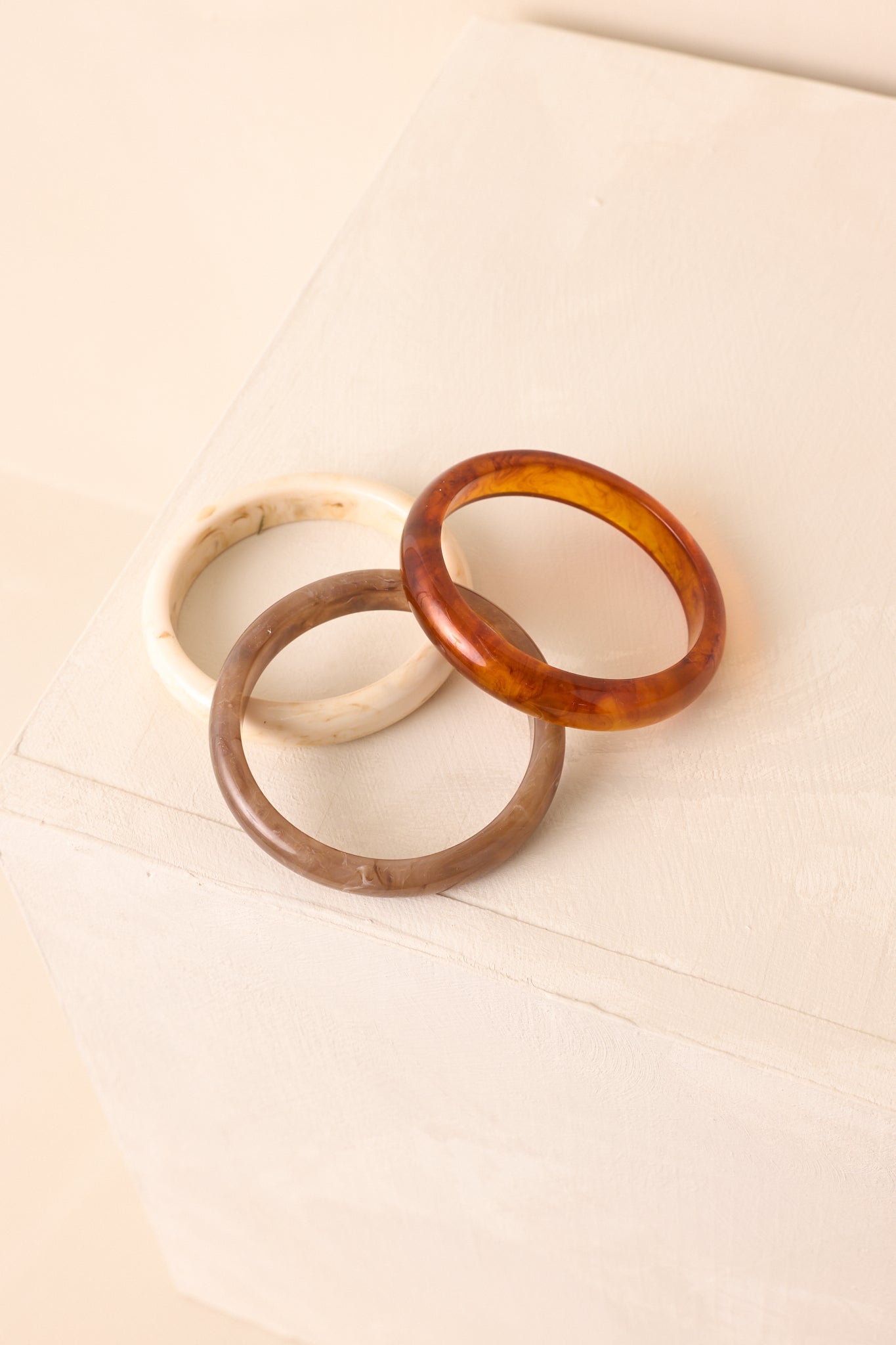 A bracelet set of three marbled bangles—tortoise shell, ivory, and taupe—displayed on a beige backdrop.