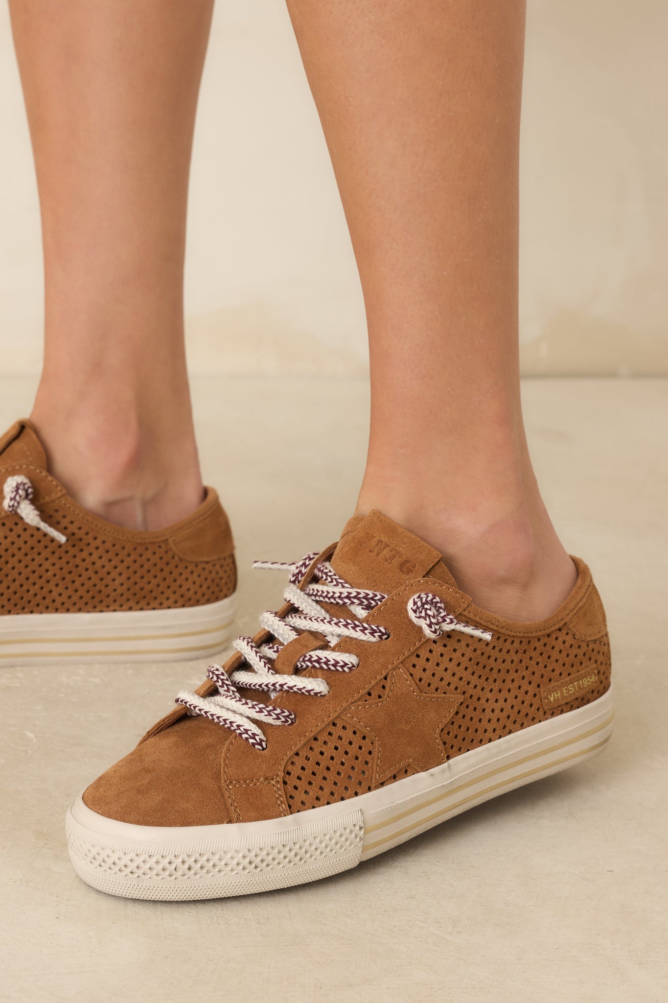 Close up of women's camel brown perforated suede lace-up sneakers with a star detail, striped white platform sole, and multicolor laces. Shown on a model from the ankle down against a neutral background.