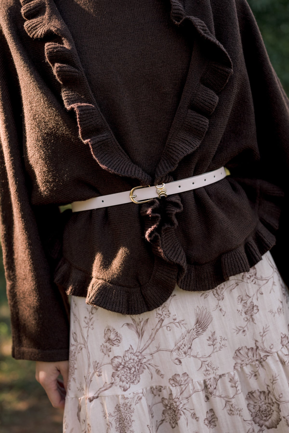 Close-up of a chocolate brown ruffle-trim cardigan cinched with a slim cream belt over a cream floral skirt.