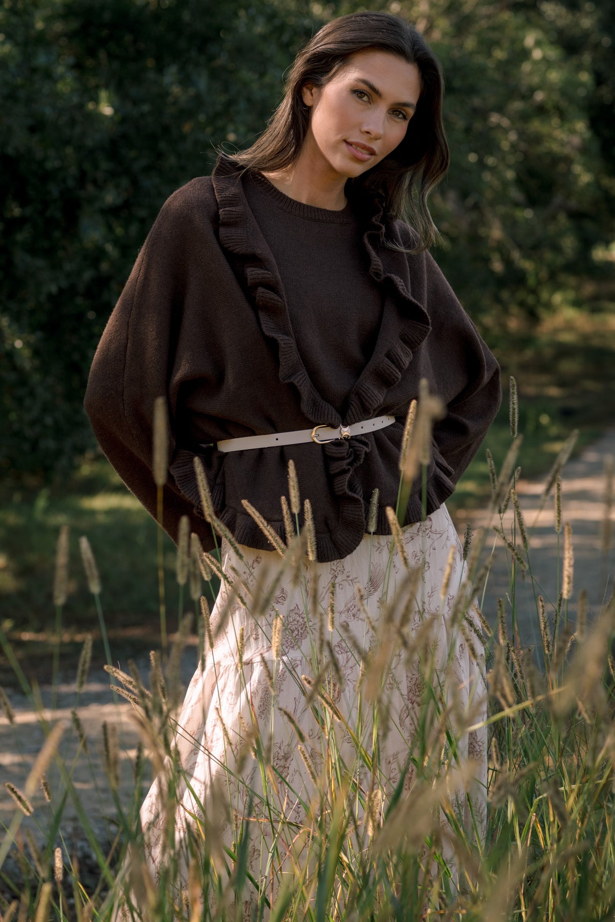 Model wearing a chocolate brown ruffle-trim top with a slim cream belt and cream floral skirt, standing among tall grasses.