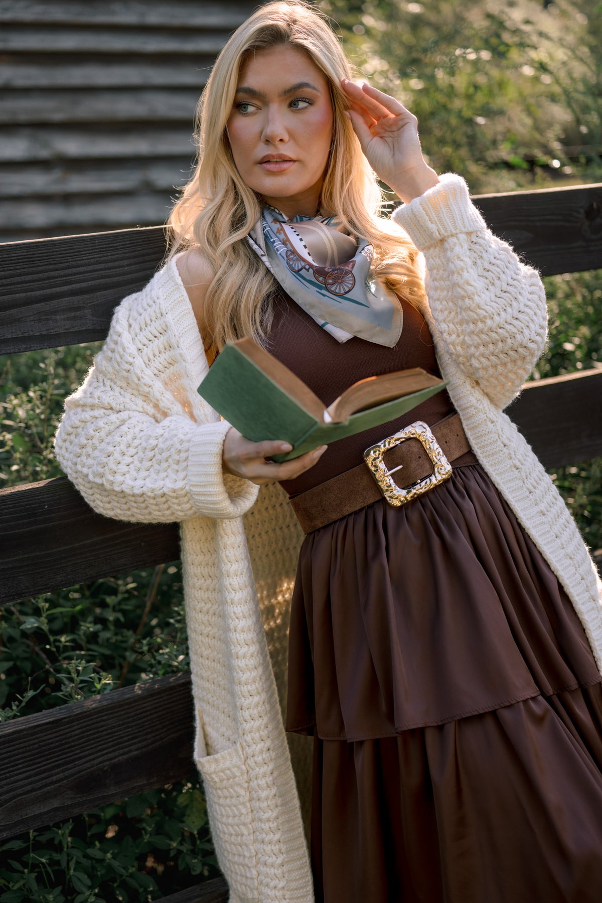 Model wearing a chocolate brown tiered dress with a wide gold-buckle belt and cream knit cardigan, holding an open book.