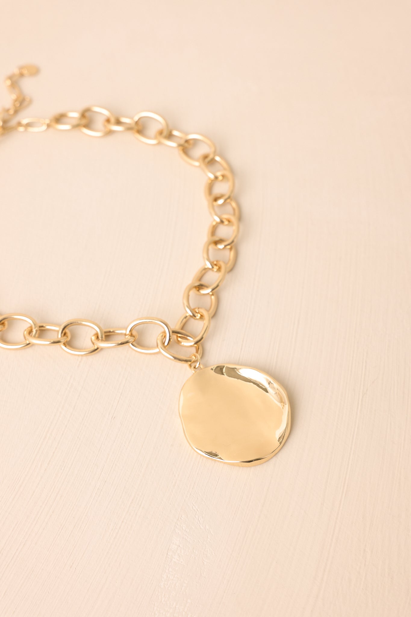 Chunky gold chain necklace with hammered organic pendant displayed on beige backdrop.