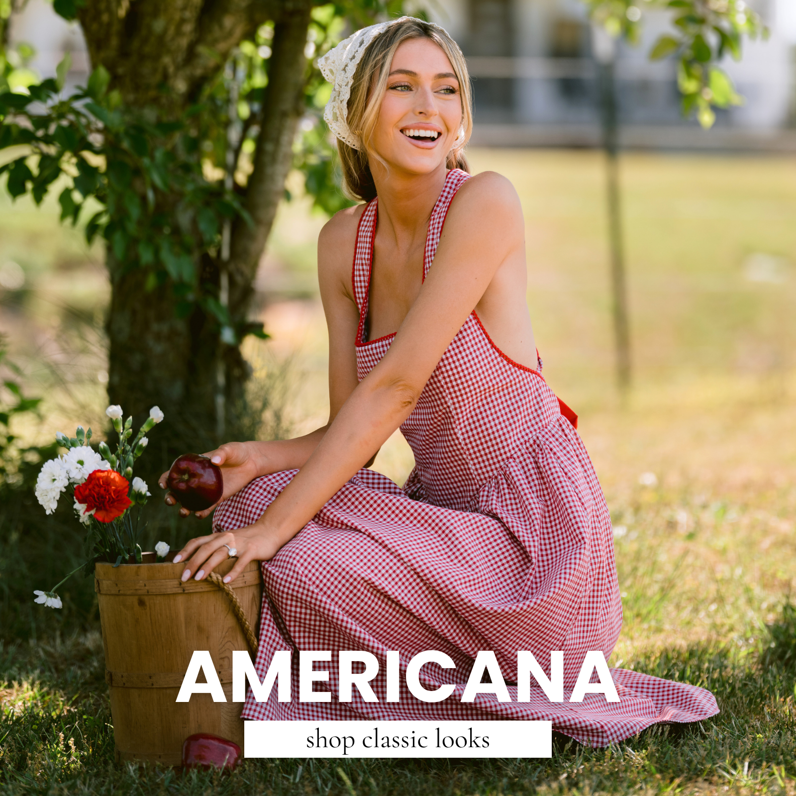 Woman wearing a red and white gingham sleeveless maxi dress with a lace headscarf, sitting beneath a tree holding a red apple beside a wooden flower basket, creating a classic Americana-inspired summer fashion scene.