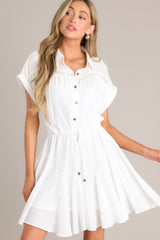 Close up front view of this white dress features a collared neckline, semi-transparent lace, a half button front, a self-tie waist feature, a lightweight fabric, and folded short sleeves.