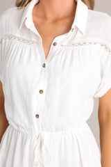 Close up view of this white dress features a collared neckline, semi-transparent lace, a half button front, a self-tie waist feature, a lightweight fabric, and folded short sleeves.