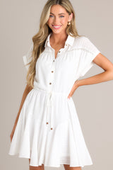 This white dress features a collared neckline, semi-transparent lace, a half button front, a self-tie waist feature, a lightweight fabric, and folded short sleeves.