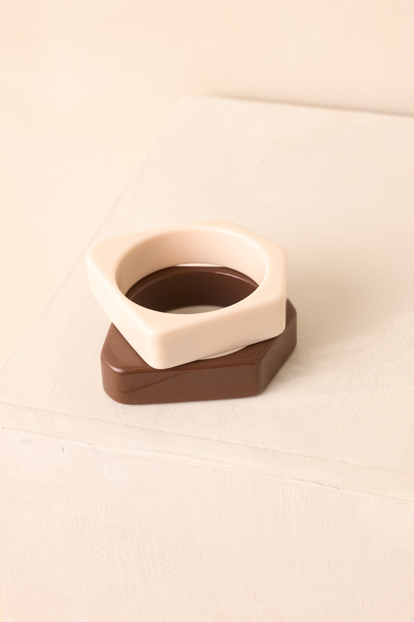 A set of two smooth, high-gloss bangles in cream and brown with square-edge design shown against a beige background.