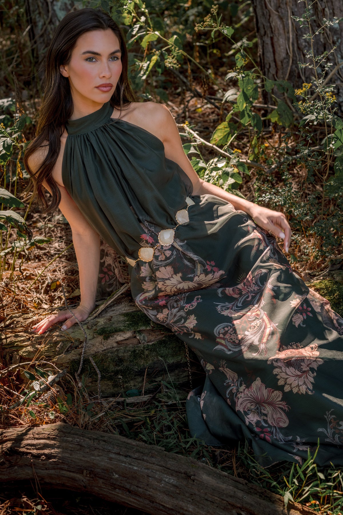 Model wearing a dark green halter maxi dress with a gathered bodice and pink floral skirt, seated on a fallen log.