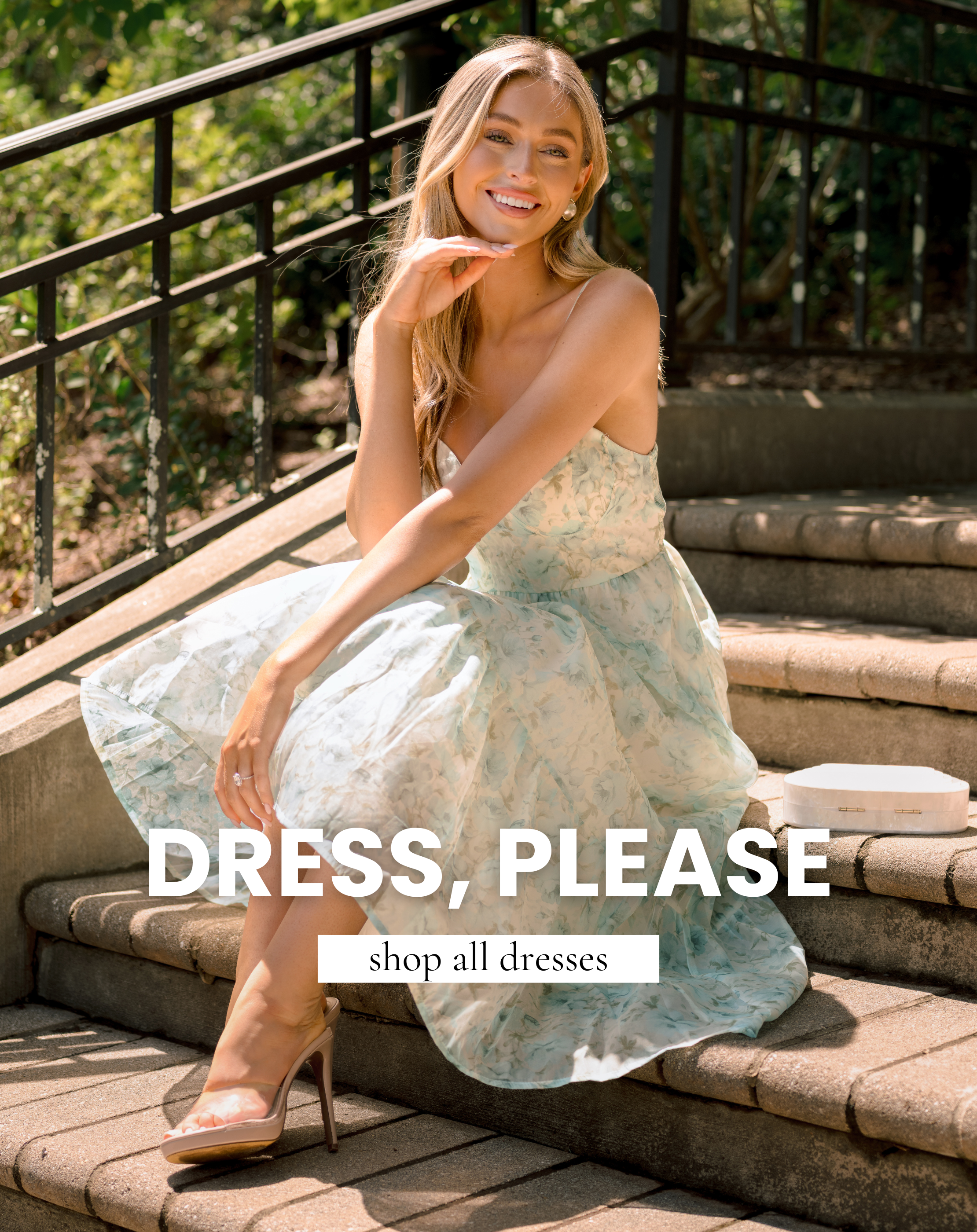 Model sitting on outdoor stone steps wearing a light blue floral maxi dress with text promoting the shop all dresses collection.