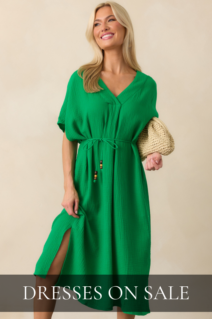 Model wearing a vibrant green midi dress with a waist tie, carrying a woven clutch and smiling in a studio setting. Dress is featured in All Dresses On Sale Shop. 