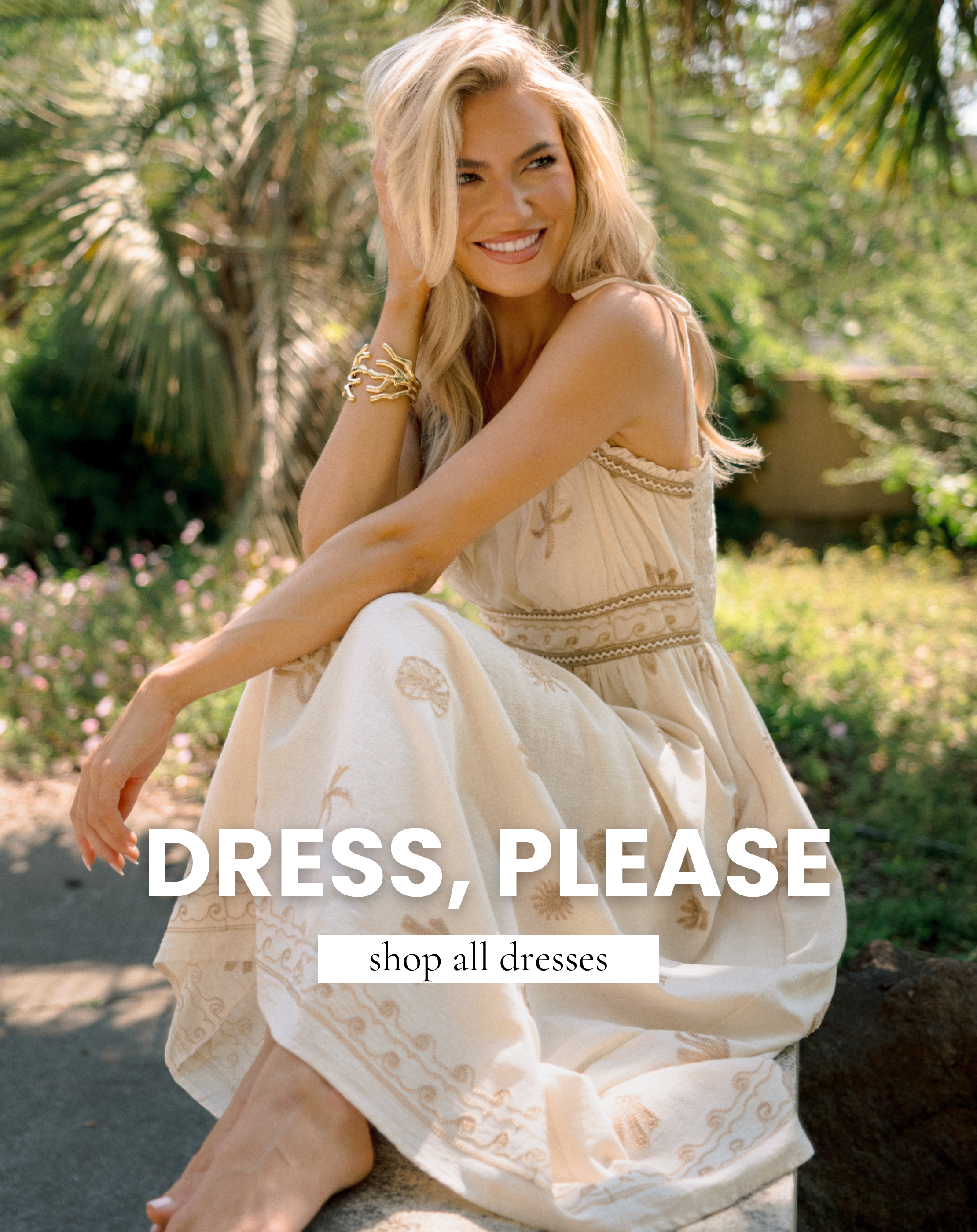 Blonde woman wearing a cream embroidered maxi dress sitting outdoors in a tropical garden setting, styled with gold bracelets for chic resort fashion inspiration.