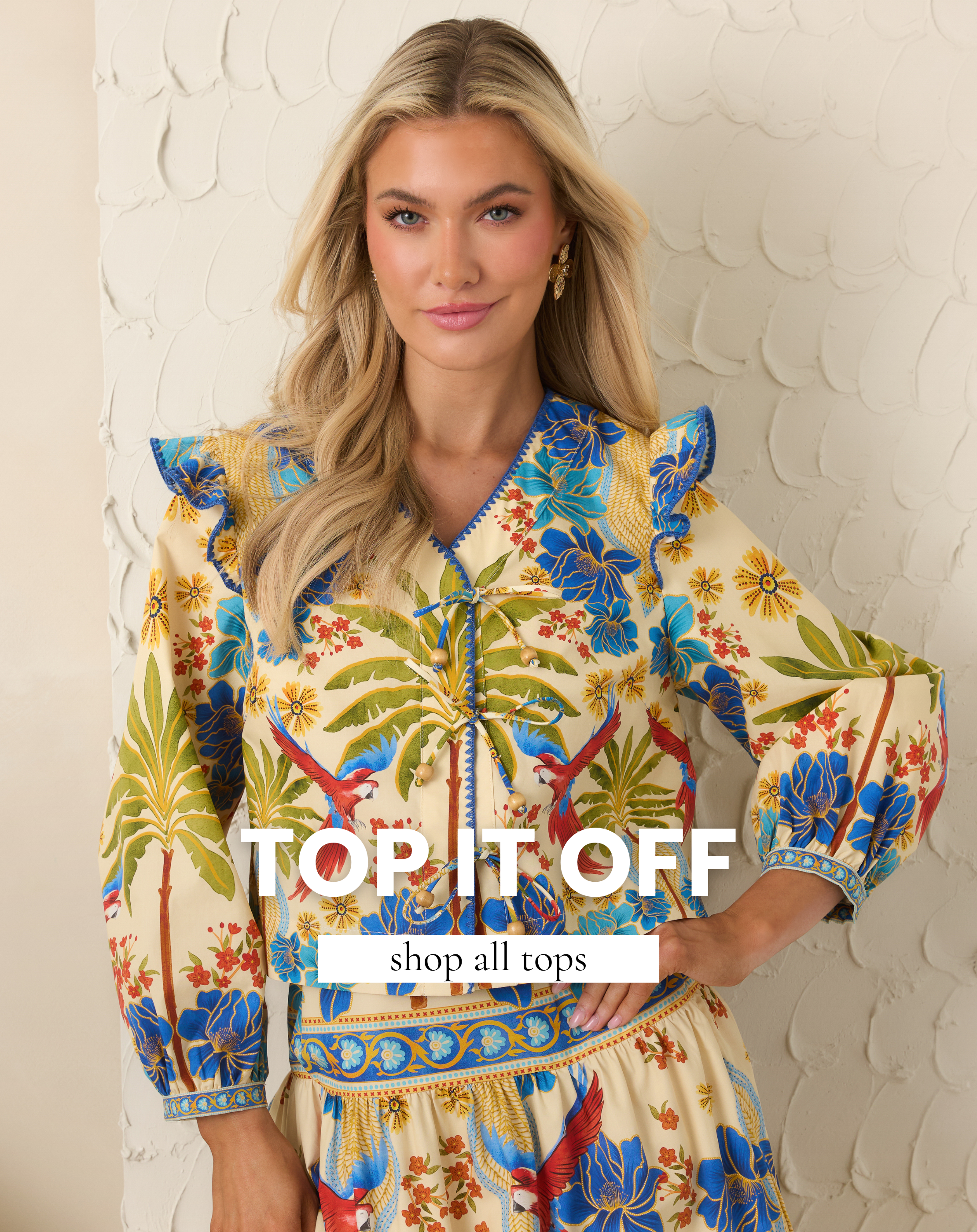 Model wearing a colorful floral and parrot print blouse with ruffle shoulders and tie front detail. This top is featured in All Tops.