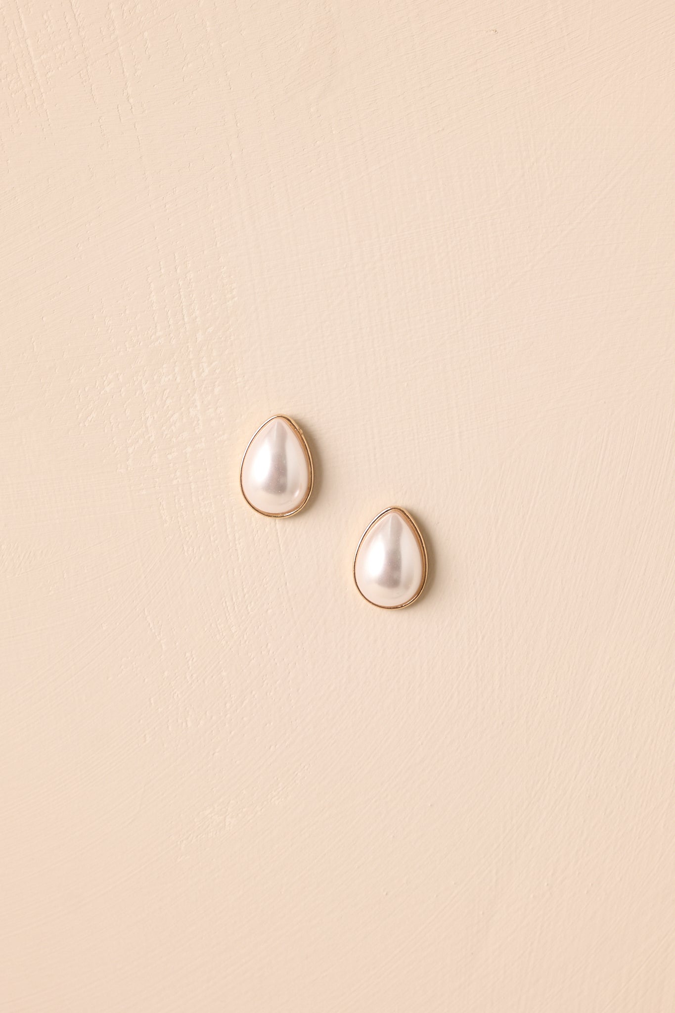 Pair of teardrop ivory pearl earrings displayed against beige background, showing gold bezel edges.