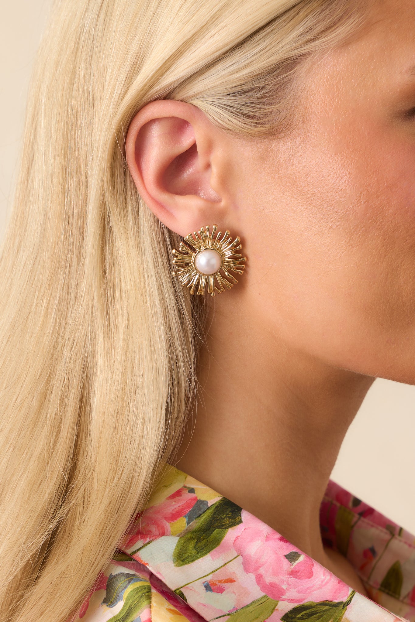 Up close photo of model wearing earrings that feature a gold textured finish, an ivory faux pearl center, surrounding sunflower petals, and secure post backings.