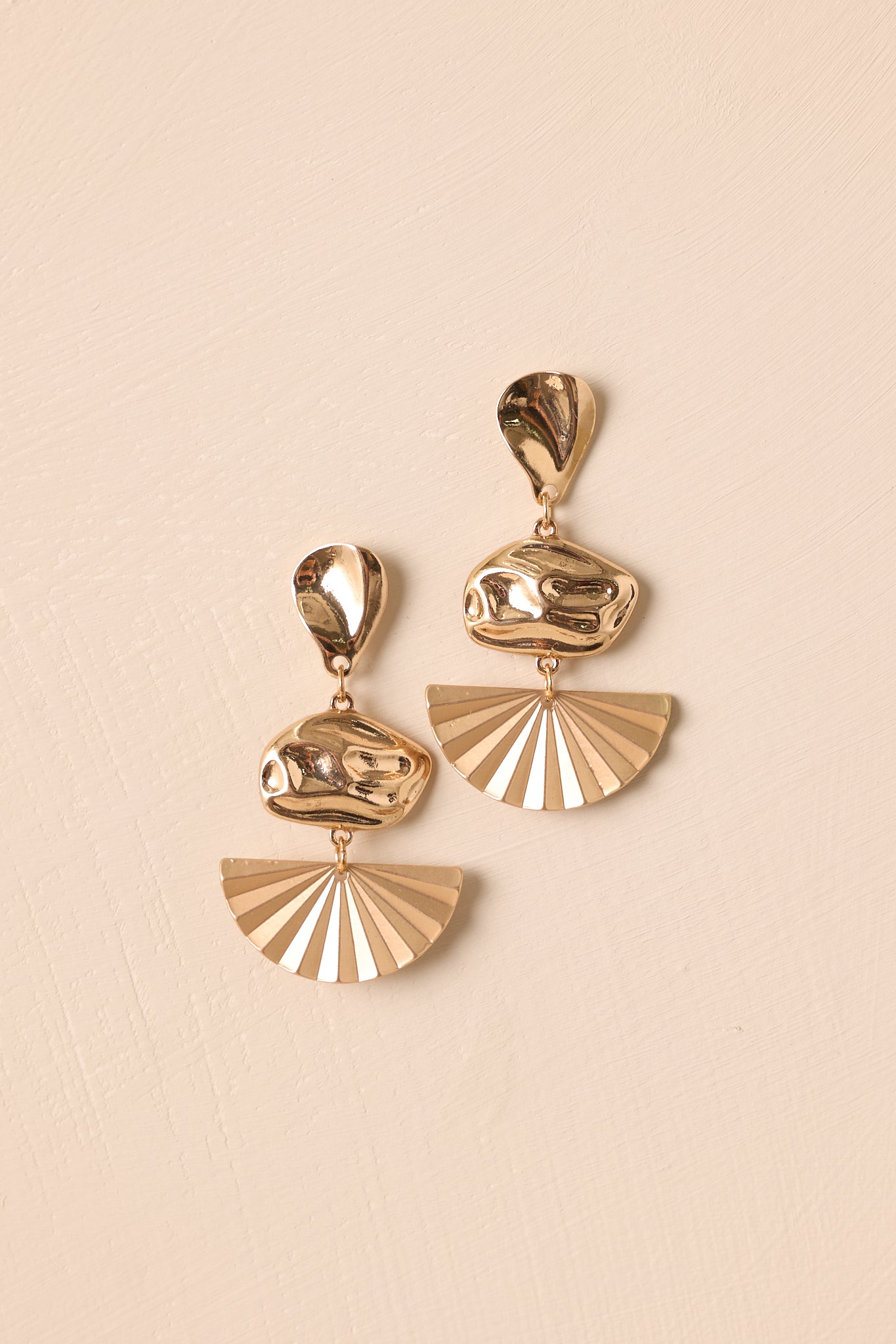 Flat lay of gold drop earrings featuring teardrop stud, oval charm, and matte textured fan charm.