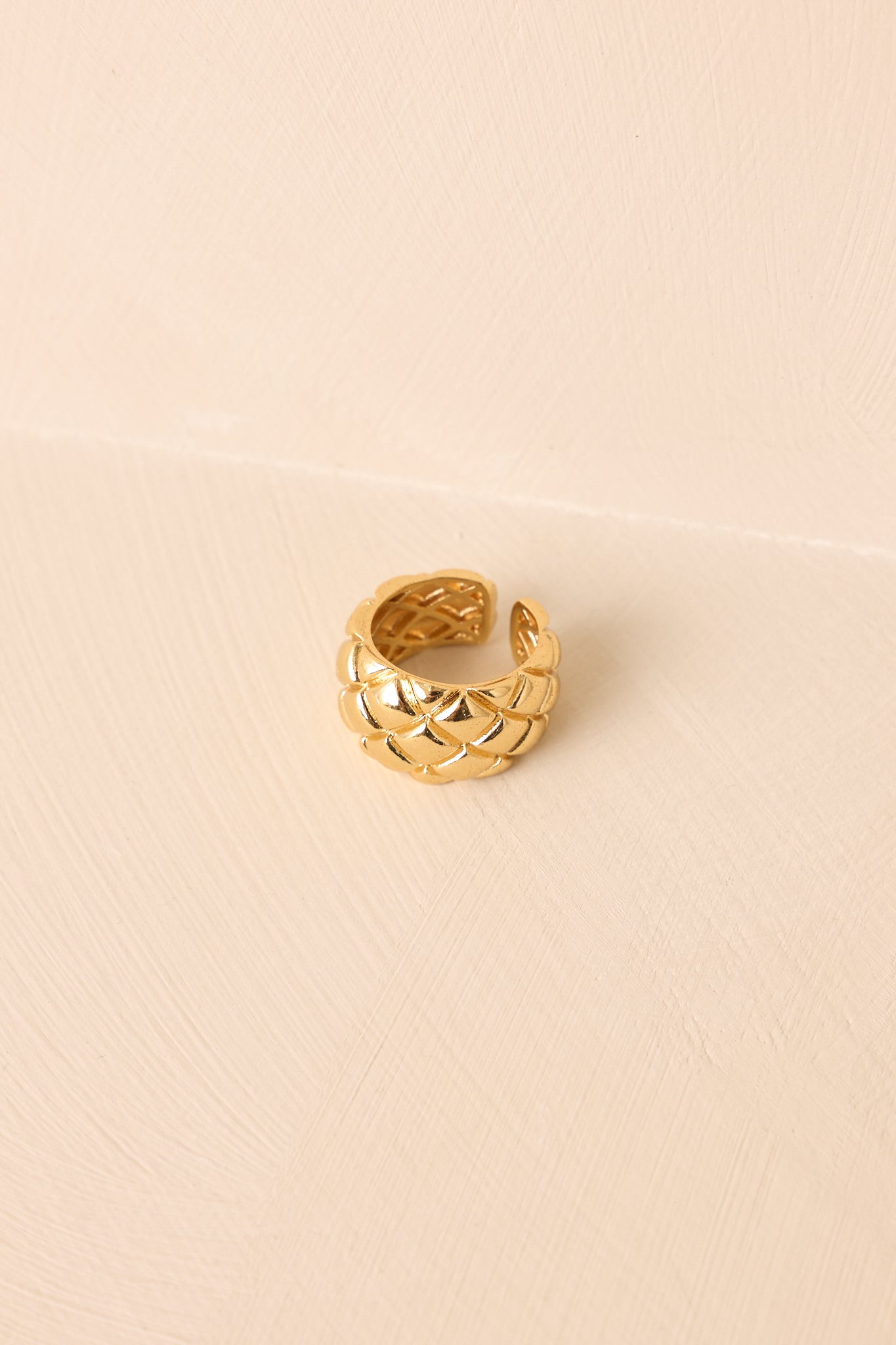 Gold ring with adjustable open cuff design and raised geometric woven texture, shown on beige background.