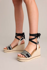 Side view of these black platform wedge sandals that feature a rounded toe, a strap over the top of the foot and around the heel, adjustable self-tie straps around the ankle, and a wedged heel.