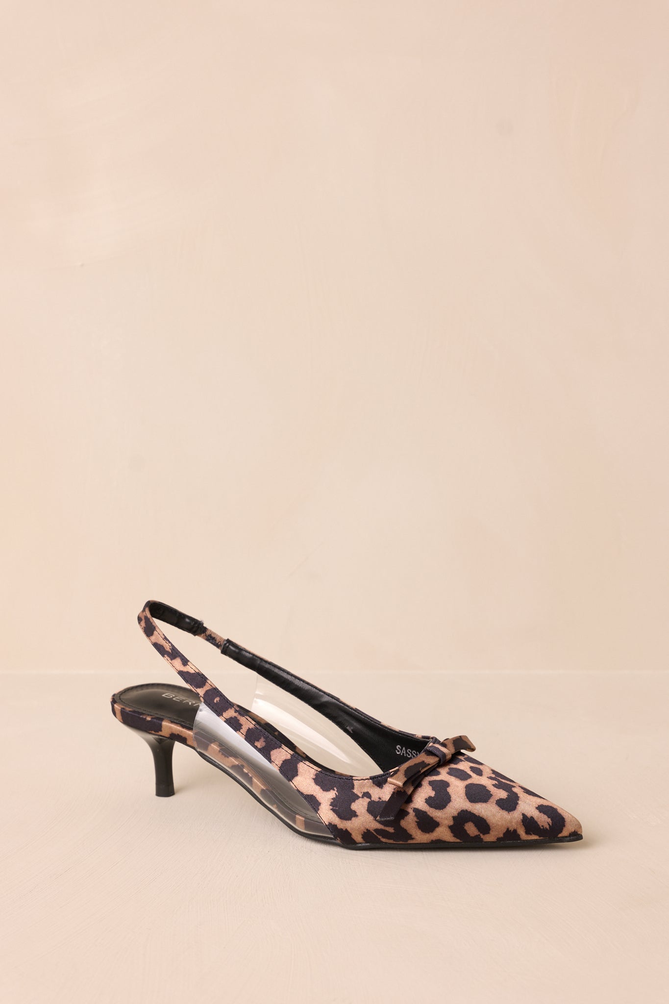 Front angled view of leopard kitten heels featuring a pointed closed toe, delicate bow detailing, a strap around the back of the foot, clear siding for extra support, and an extremely short heel
